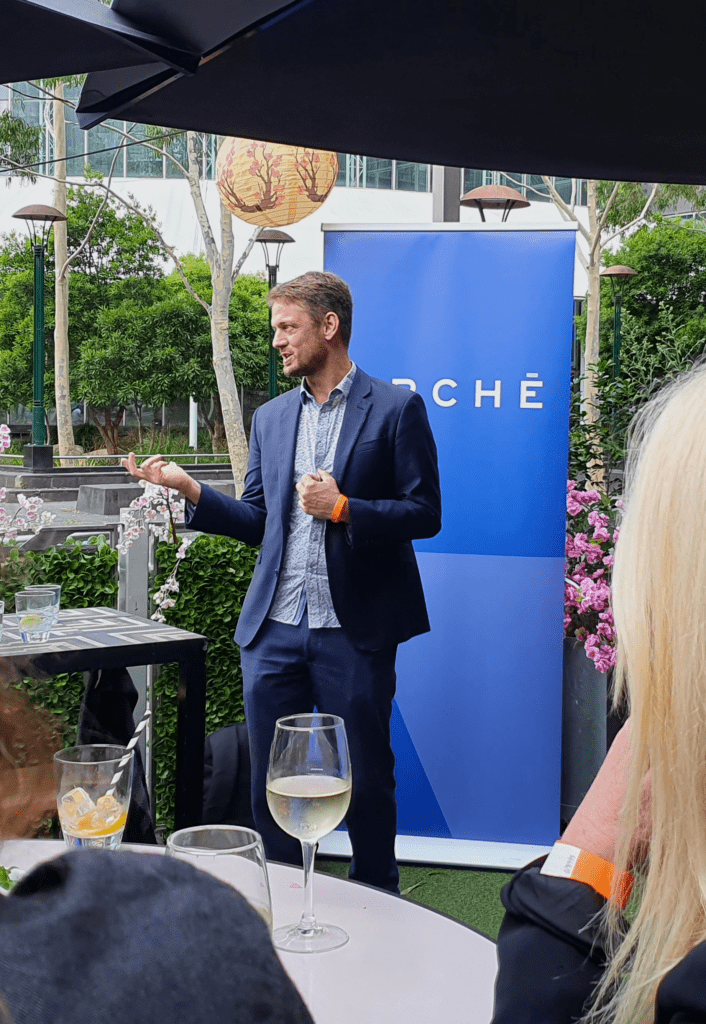 Andrew Murdoch mid-speech at a casual outdoor bar, standing in front of Arche's pull-up banner
