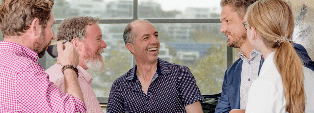 Alex Ross in the centre of a cheerful business casual group in front of a big, bright window that looks out on a leafy suburb. On his right are Peter Bradnam and Matt Richards; on his left are Andrew Murdoch and Ava Murdoch. 